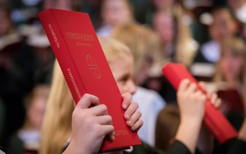 Två personer håller upp kyrkohandboken i luften. En flickkör syns suddigt i bakgrunden.