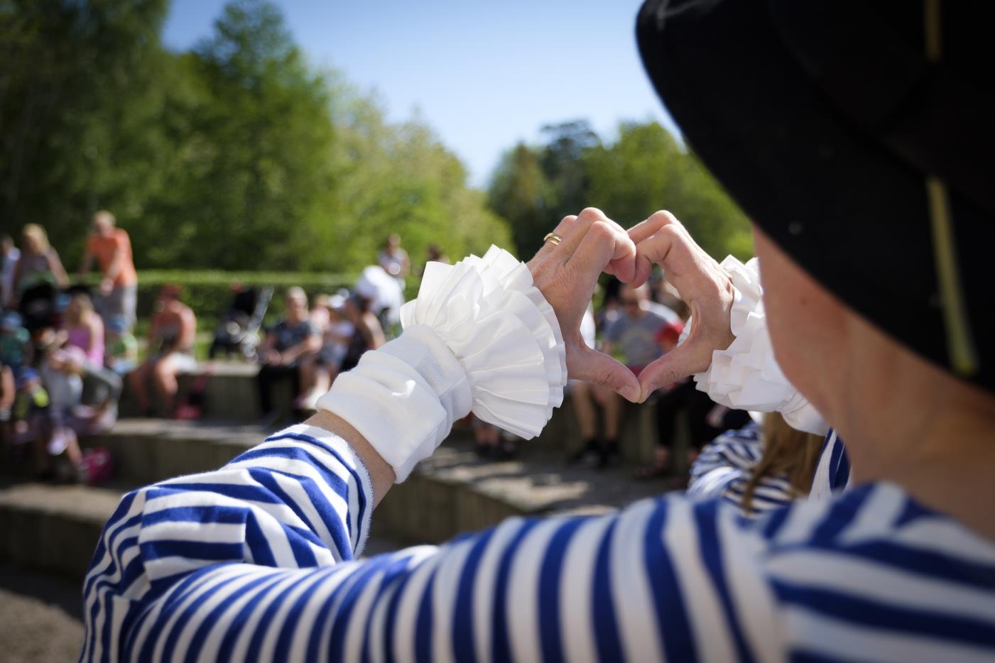 En person i blåvitrandig tröja och svart hatt gör ett hjärta med händerna på en utomhusteater.
