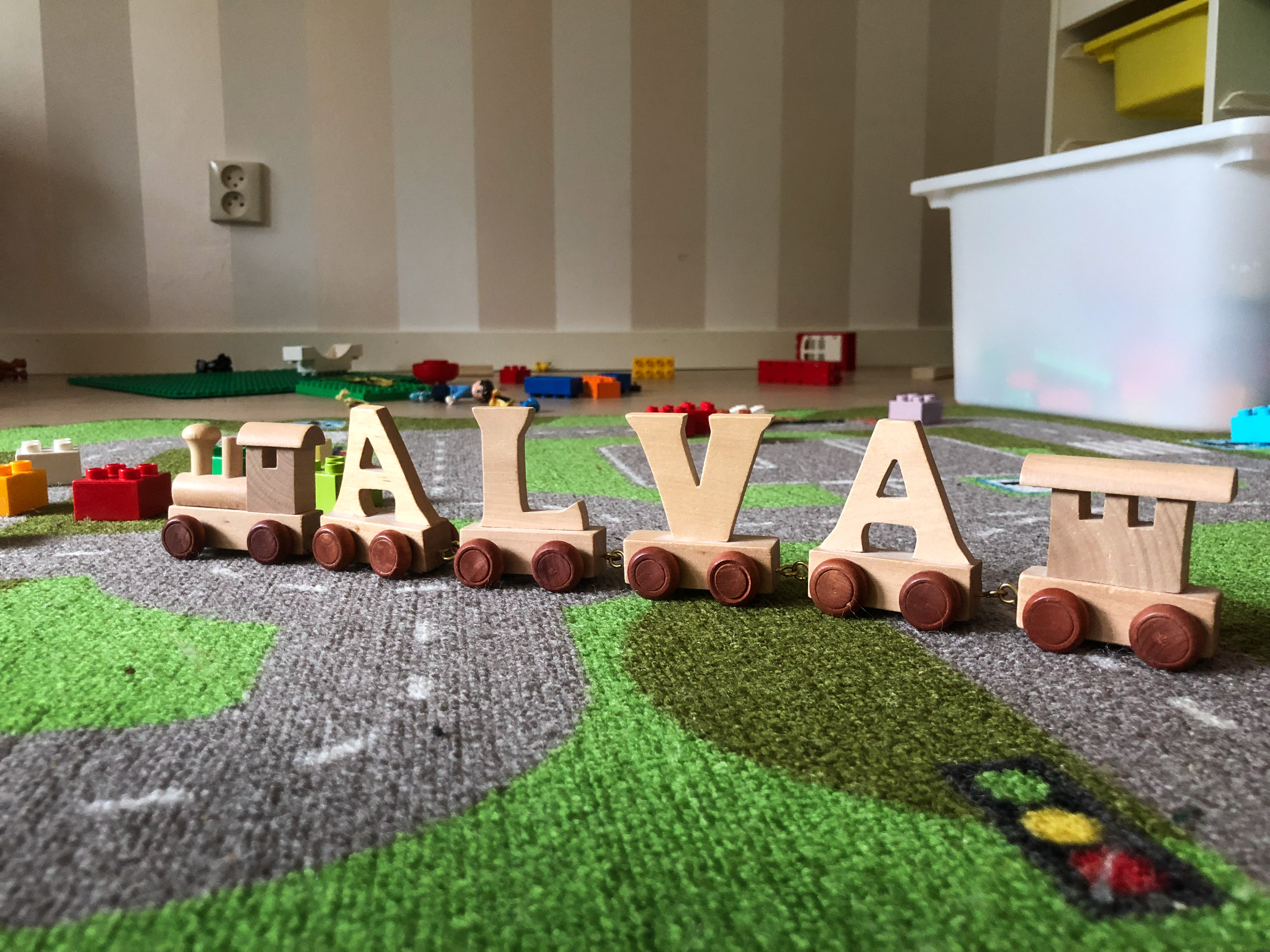 Ett leksakståg med bokstäver på vagnarna som bildar namnet Alva står på golvet i ett lekrum.