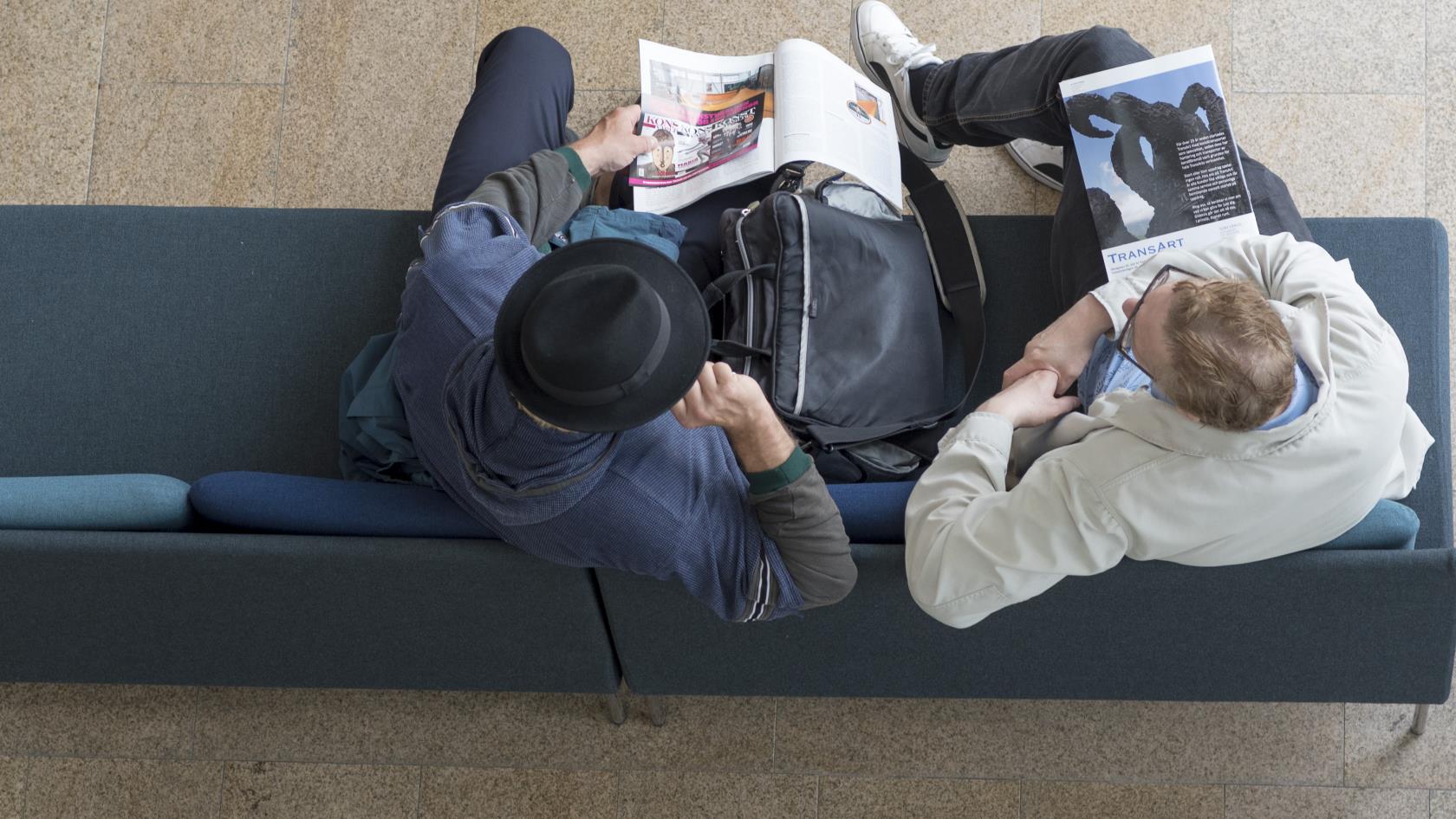 Två män fotograferade ovanifrån sitter i en soffa med varsin tidning.