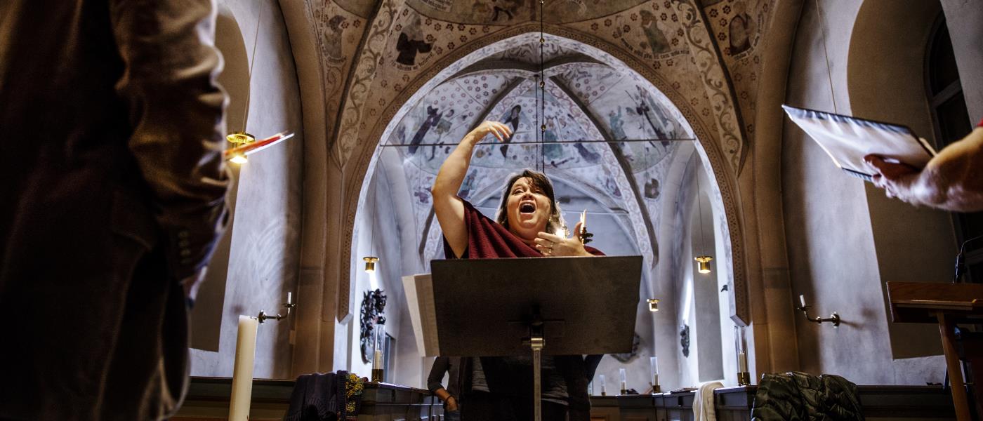 En kvinnlig körledare dirigerar i en kyrka. Några körmedlemmar syns suddigt i förgrunden.