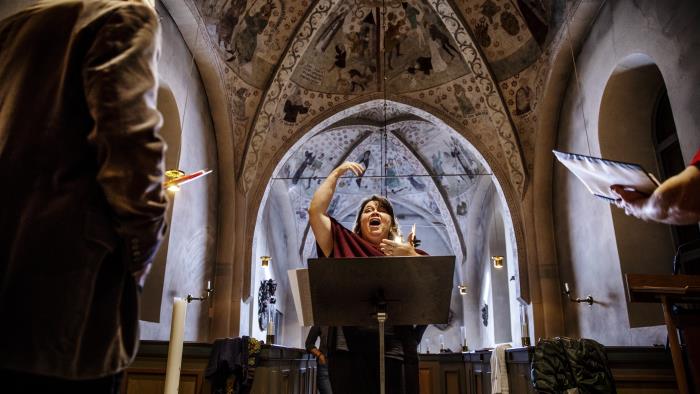 En kvinnlig körledare dirigerar i en kyrka. Några körmedlemmar syns suddigt i förgrunden.