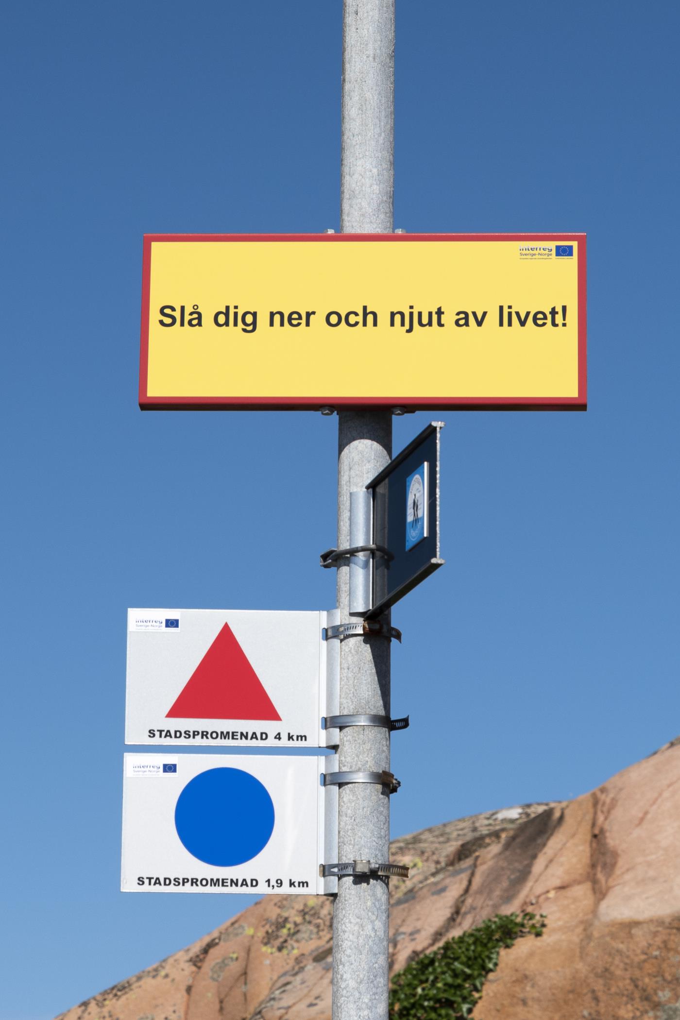 En gul skylt på en stolpe med texten Slå dig ner och njut av livet.
