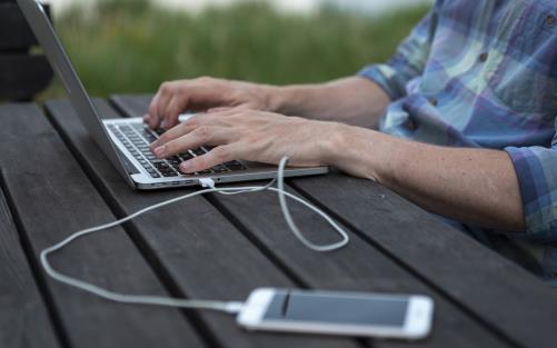 En person sitter utomhus och arbetar på sin laptop. En mobiltelefon ligger bredvid kopplad med en sladd till datorn.