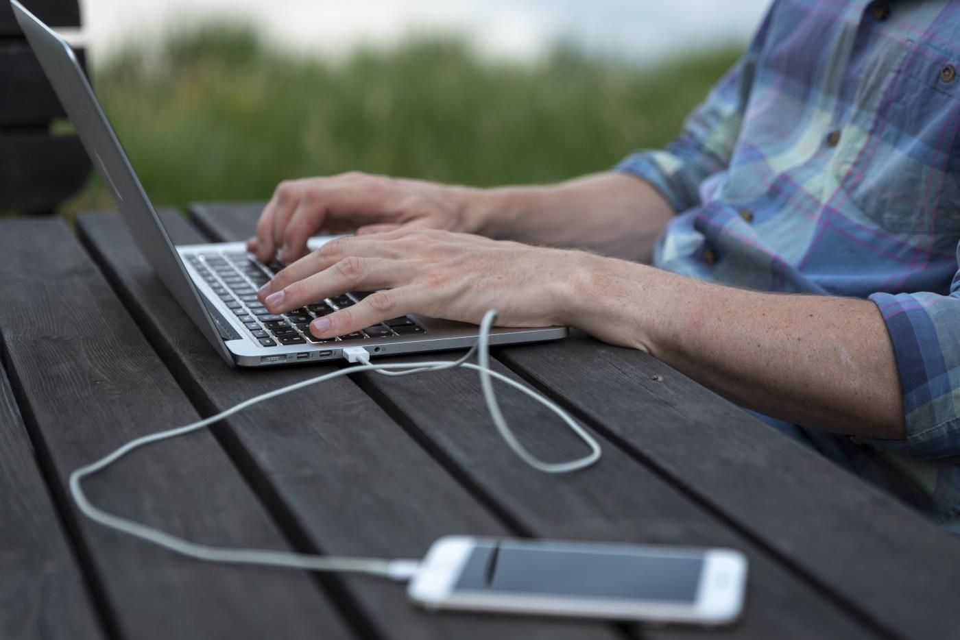 En person sitter utomhus och arbetar på sin laptop. En mobiltelefon ligger bredvid kopplad med en sladd till datorn.
