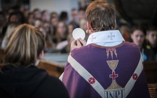 En präst med lila mantel håller upp en oblat i en fullsatt kyrka.