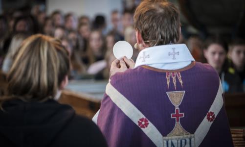 En präst med lila mantel håller upp en oblat i en fullsatt kyrka.