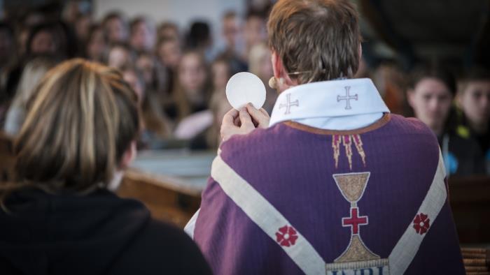 En präst med lila mantel håller upp en oblat i en fullsatt kyrka.