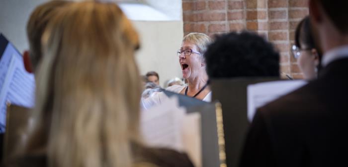En kvinnlig körledare står i en kyrka och dirigerar. Körmedlemmar i förgrunden.