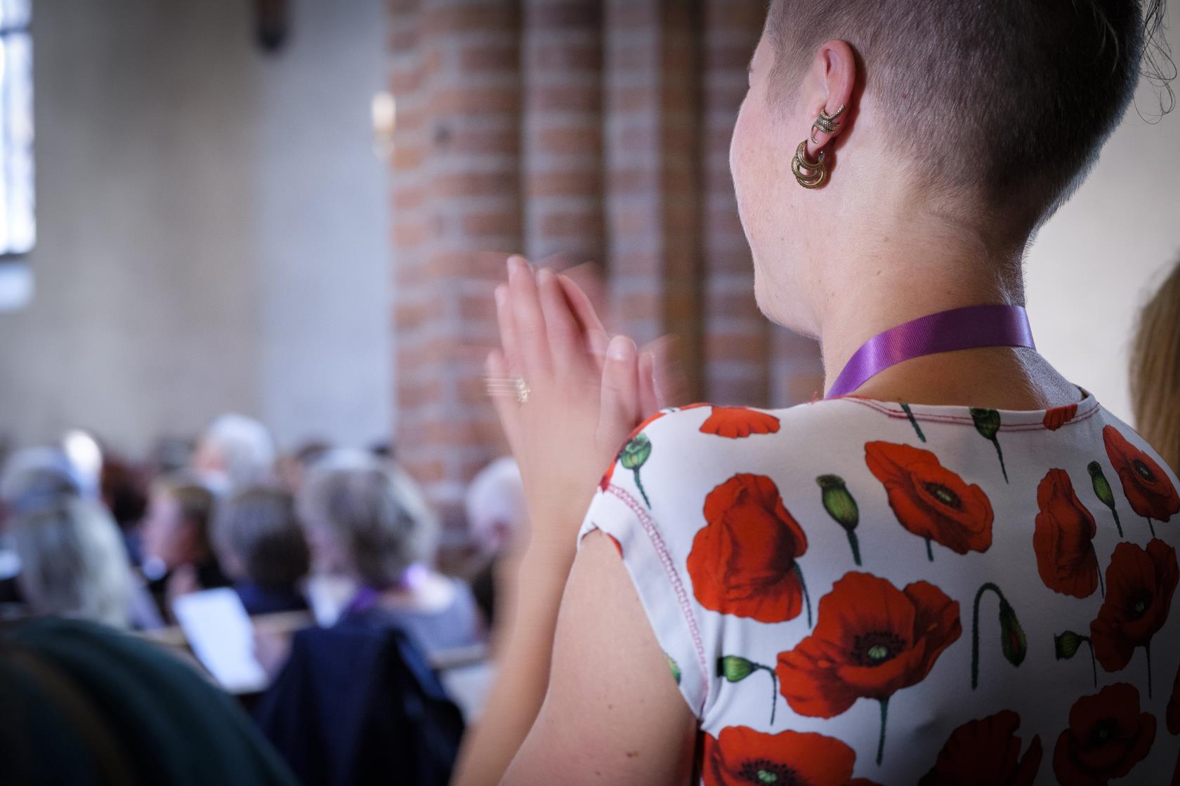 Närbild på en kvinna som står upp med knäppta händer i en kyrka. Fyllda kyrkbänkar syns suddigt i bakgrunden.