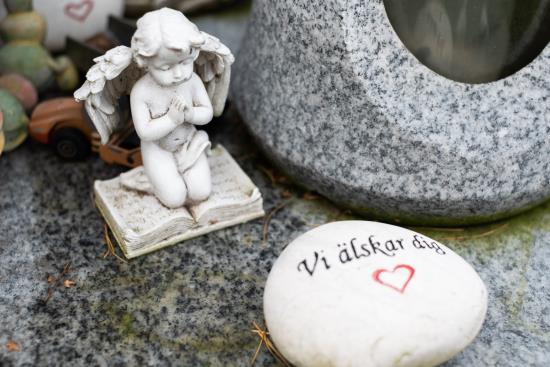 Ett vitt stenhjärta med texten Vi älskar dig och en ängel på knä ligger vid en grav.