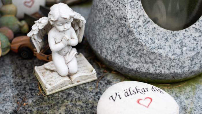 Ett vitt stenhjärta med texten Vi älskar dig och en ängel på knä ligger vid en grav.