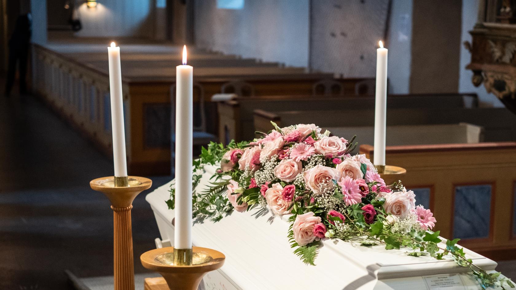 En vit kista med rosa blommor står omringad av ljus uppe vid koret.