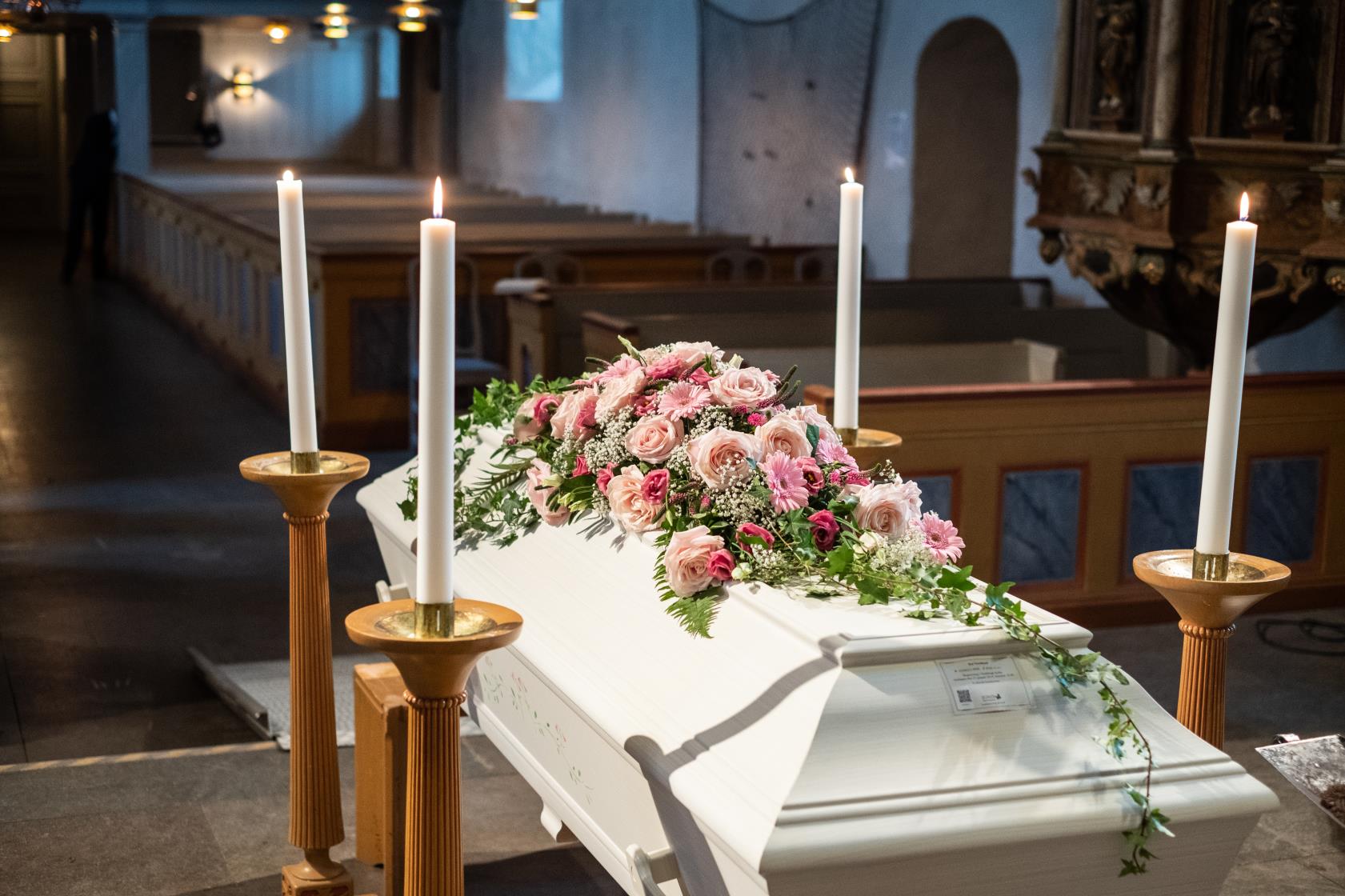 En vit kista med rosa blommor står omringad av ljus uppe vid koret.