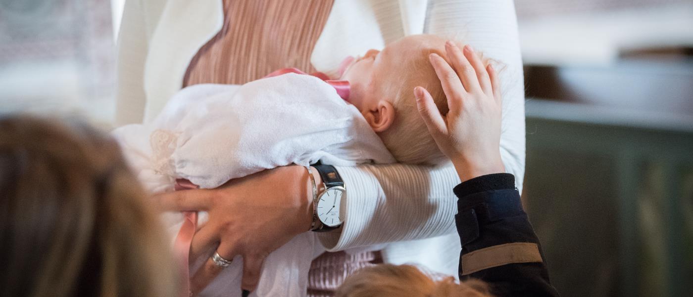 En bebis i dopklänning sover i någons famn. Ett barn klappar på bebisens huvud.