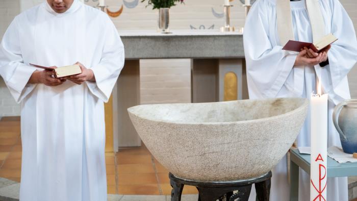 En präst och en man i vit dopkåpa står på varsin sida om en dopfunt.
