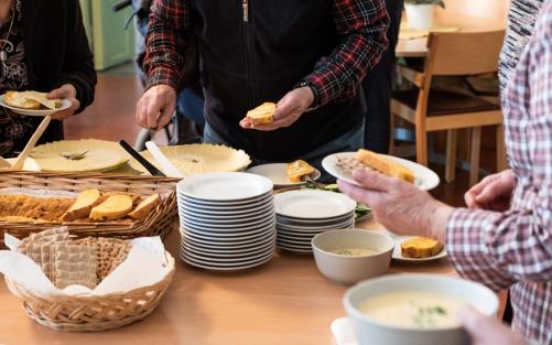 Runt ett fikabord står några personer och brer sina smörgåsar.