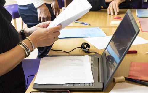 På ett mötesbord står en bärbar dator uppslagen. Papper och pennor ligger utspridda framför mötesdeltagarna.