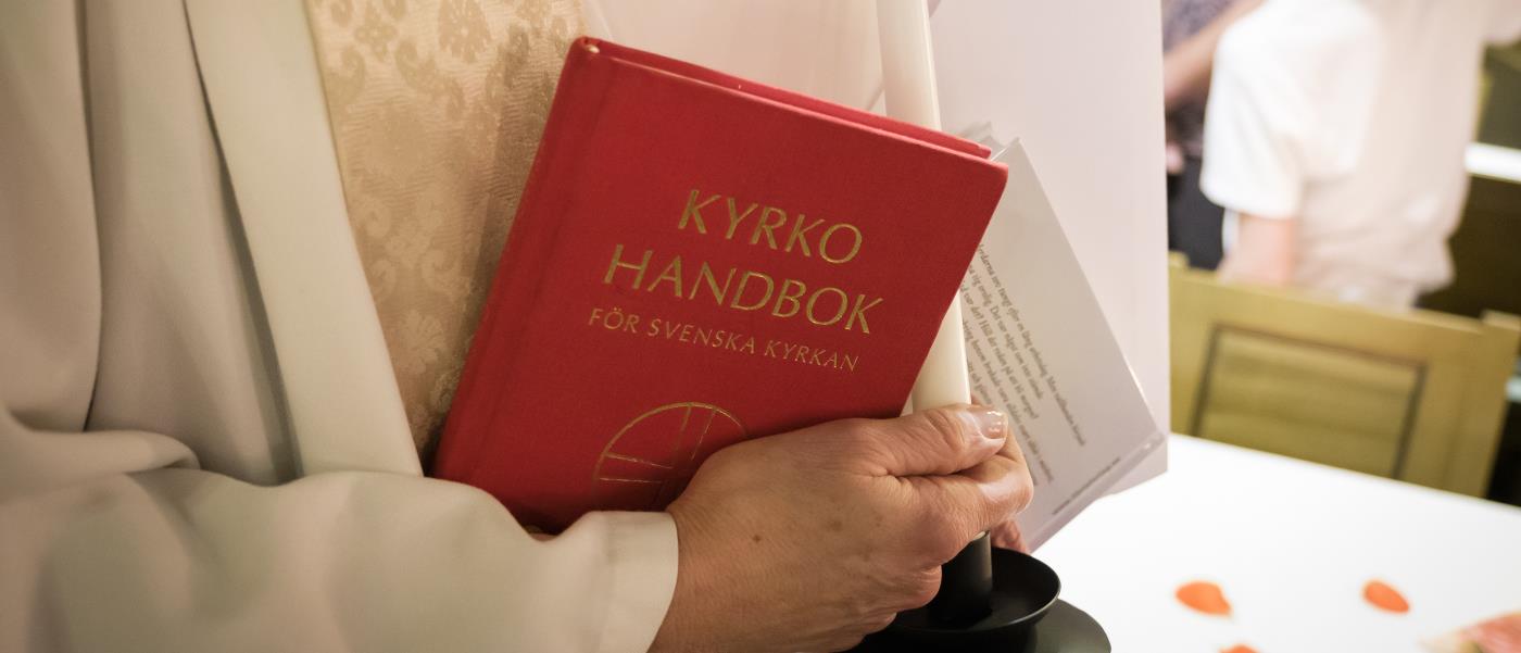 En präst håller ett ljus, ett vitt kuvert och en kyrkohandbok i händerna.