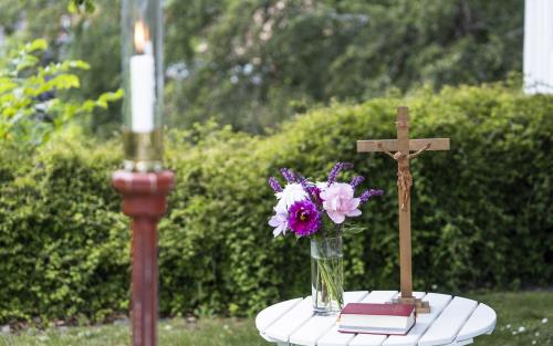 Ett litet bord med ett krucifix och en blombukett står i en trädgård. En ljusstake i förgrunden.
