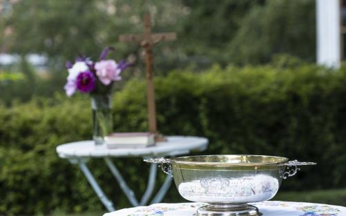 Ett bord med en liten dopfunt står i trädgården. Längre bort står ett till bord med ett krucifix och en blombukett.