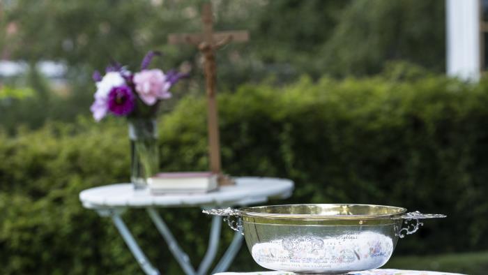 Ett bord med en liten dopfunt står i trädgården. Längre bort står ett till bord med ett krucifix och en blombukett.