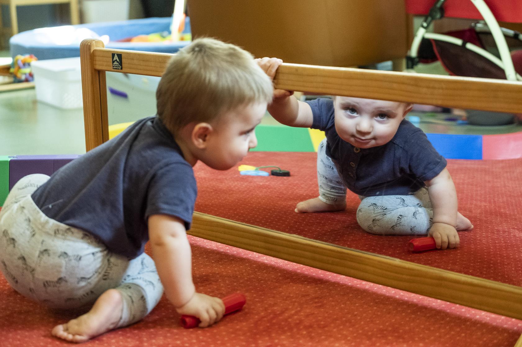 En bebis sitter på golvet och tittar in i kameran genom spegeln framför honom.