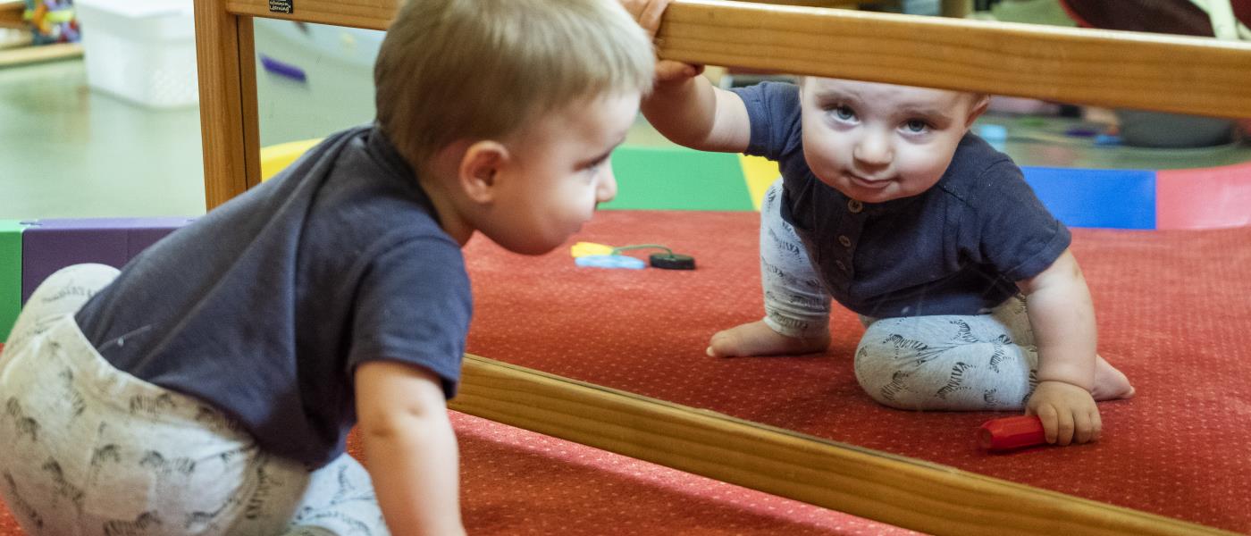 En bebis sitter på golvet och tittar in i kameran genom spegeln framför honom.
