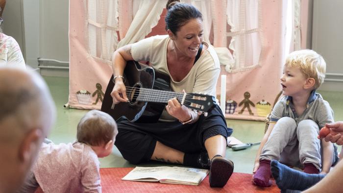 En kvinnlig ledare spelar gitarr och sjunger för en grupp barn i ring.