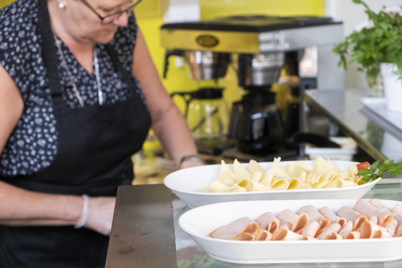 I förgrunden står två fat med upplagda smörgåspålägg på en glasdisk. Längre bort arbetar en kvinna i ett kök.