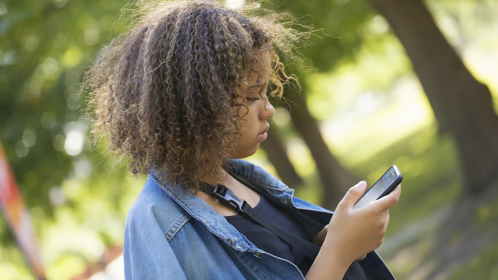 En ung tjej med jeansjacka står utomhus och tittar ner i mobiltelefonen.