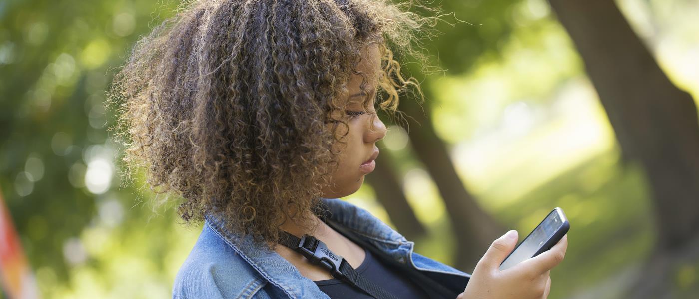En ung tjej med jeansjacka står utomhus och tittar ner i mobiltelefonen.