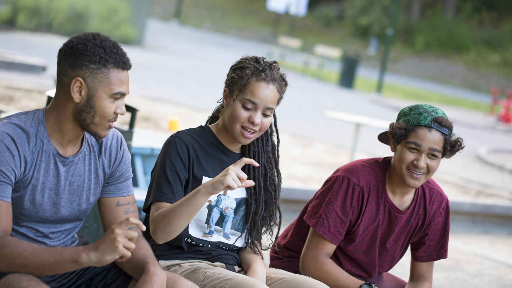 Tre ungdomar sitter utomhus i en park och gör tecken med fingrarna.