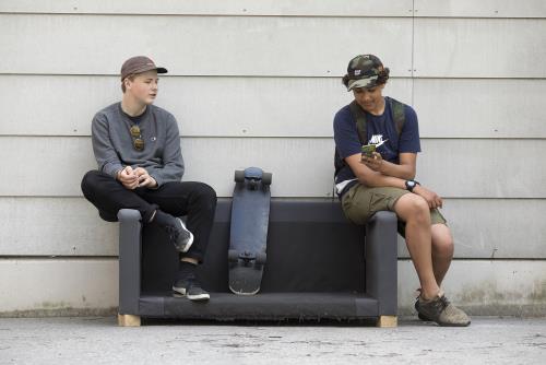 Två killar med keps och skateboard sitter utomhus och snackar på en gammal soffa.