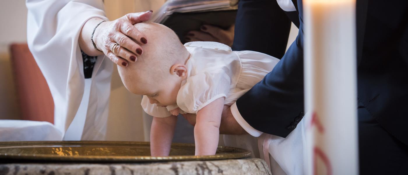 Någon sänker en bebis ner i dopfunten medan prästen håller sin hand på barnets huvud.