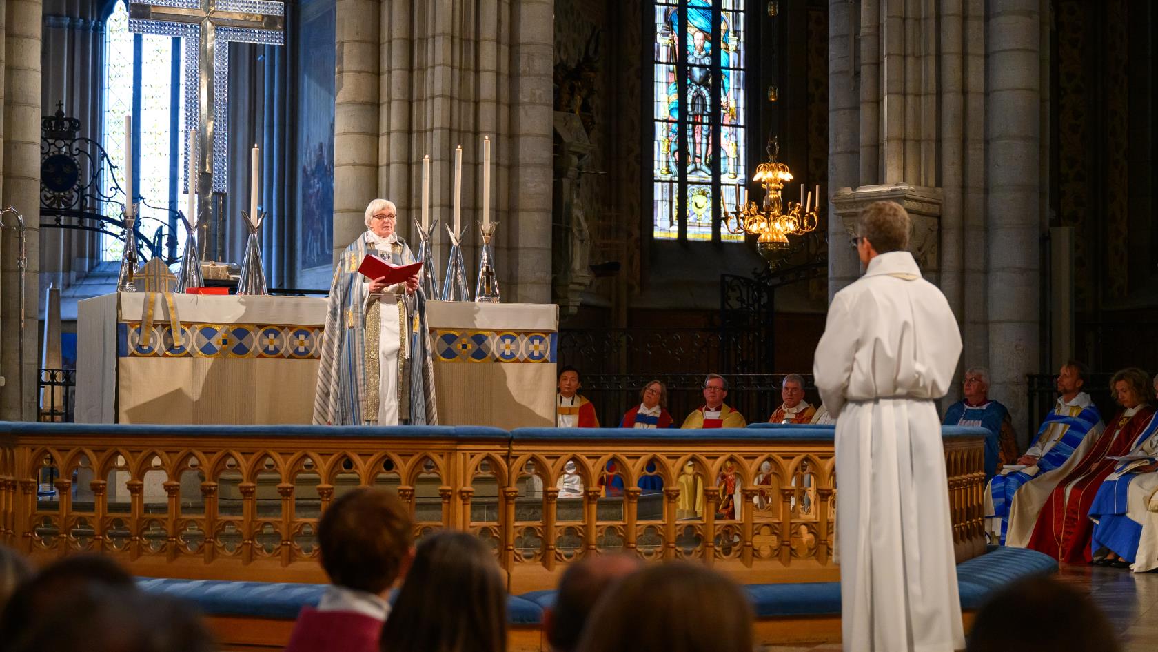 Ärkebiskop Antje Jackélen står vid altaret i Uppsala domkyrka och läser ur en bok. Framför altaret står en man i vit klädnad.