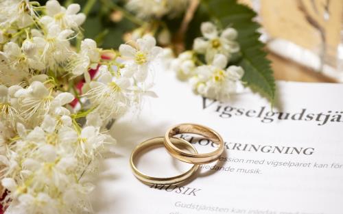 Närbild på två vigselringar som tillsammans med en blomkvist ligger på en uppslagen bok med rubriken Vigselgudstjänst.