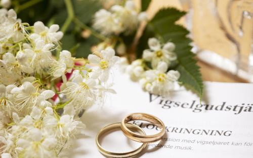 Närbild på två vigselringar som tillsammans med en blomkvist ligger på en uppslagen bok med rubriken Vigselgudstjänst.