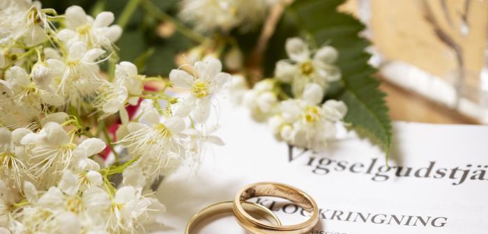 Närbild på två vigselringar som tillsammans med en blomkvist ligger på en uppslagen bok med rubriken Vigselgudstjänst.