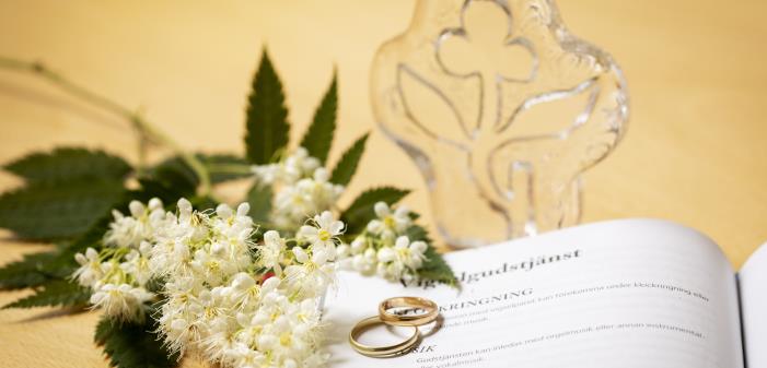 Närbild på ett par ringar och en blomkvist som ligger på en bok med rubriken Vigselgudstjänst uppslagen.