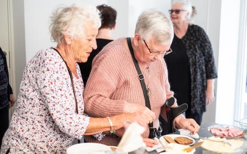 Två äldre damer gör mackor vid ett bord med olika pålägg.