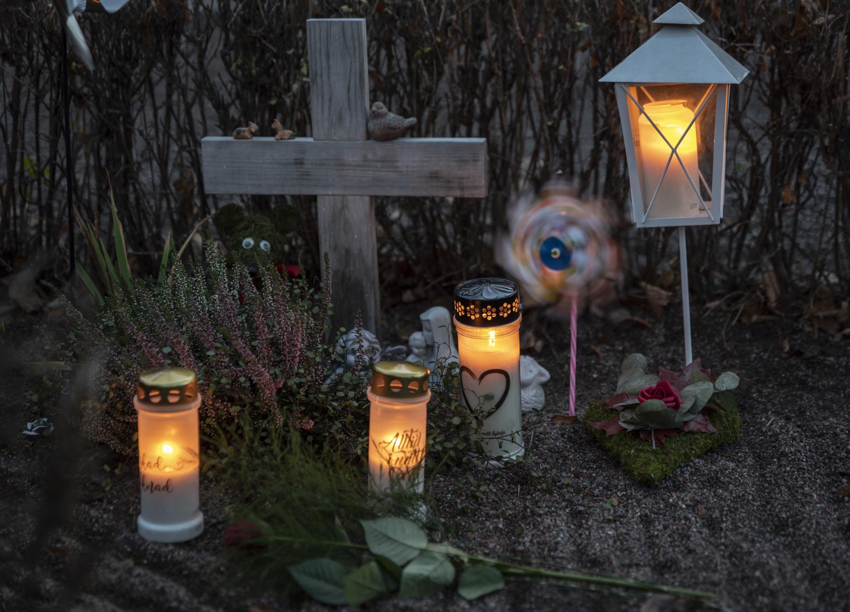 En grav med ett enkelt träkors har smyckats med gravljus, en lykta, en vindsnurra, ler- och gipsfigurer samt olika blommor.