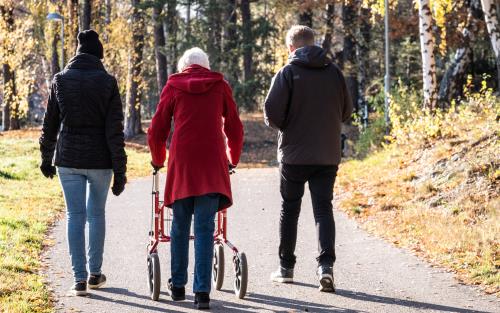 Tre personer promenerar på en asfalterad väg. Det är höst. Personerna går från oss. En har rollator.