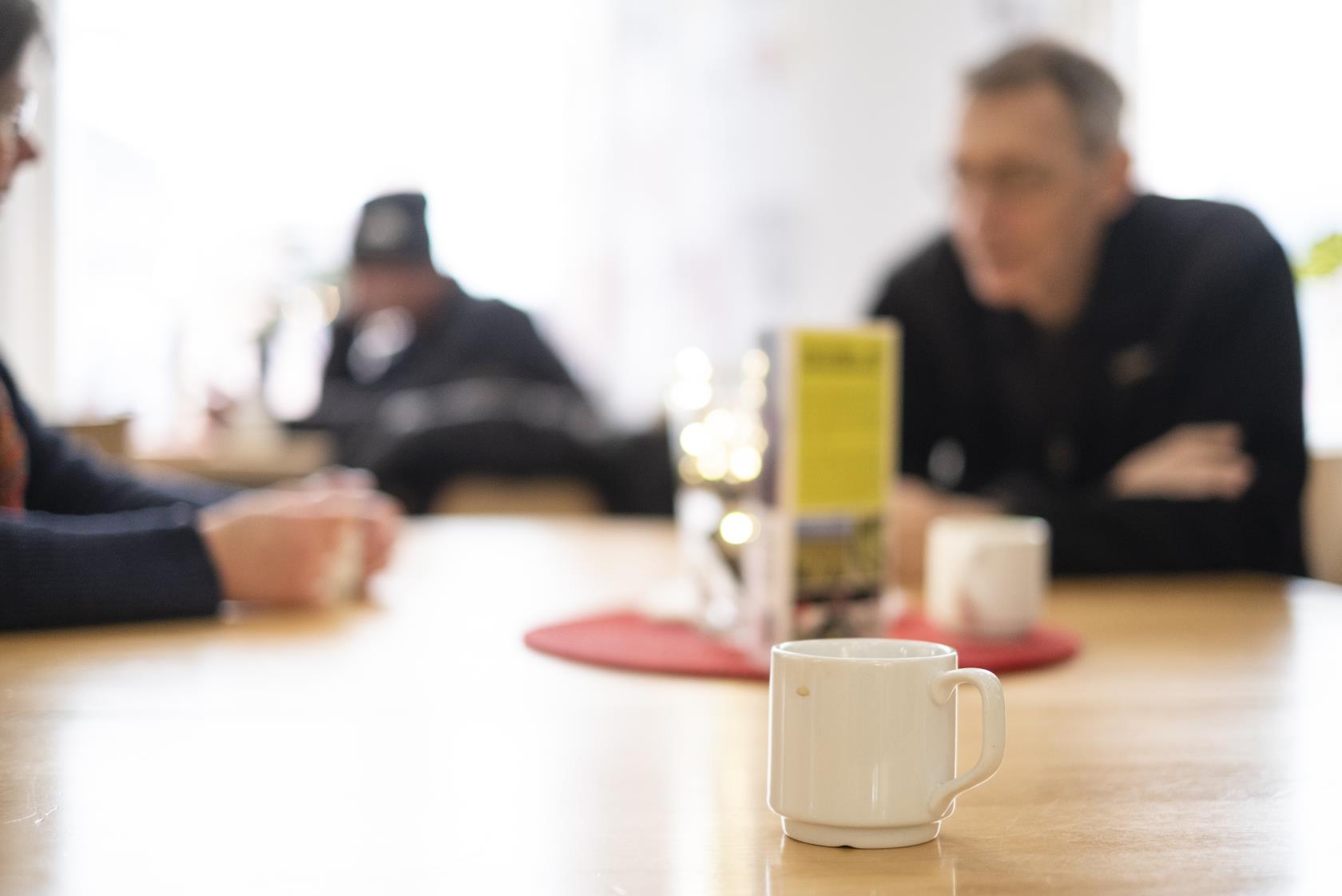 En kopp kaffe står på ett bord. I bakgrunden ses människor som samtalar.