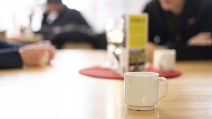 En kopp kaffe står på ett bord. I bakgrunden ses människor som samtalar.