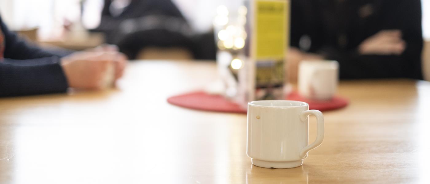 En kopp kaffe står på ett bord. I bakgrunden ses människor som samtalar.