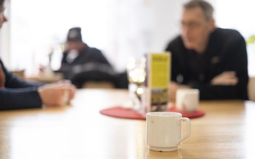 En kopp kaffe står på ett bord. I bakgrunden ses människor som samtalar.