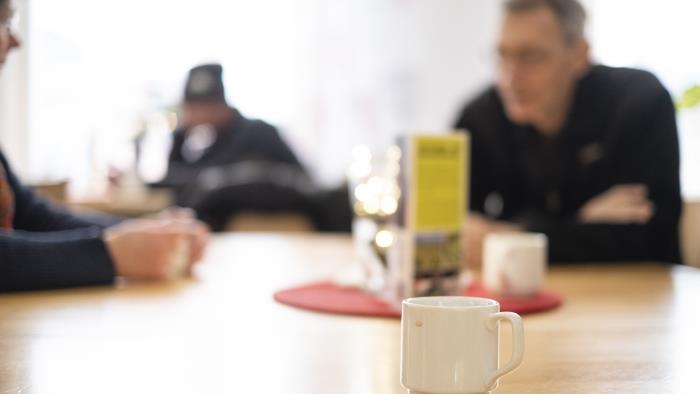 En kopp kaffe står på ett bord. I bakgrunden ses människor som samtalar.