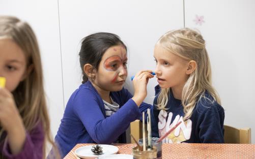 En liten flicka med ansiktsmålning målar ansiktet på en annan tjej med en penna.