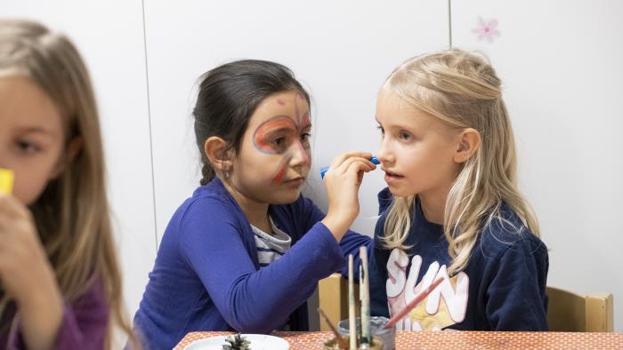 En liten flicka med ansiktsmålning målar ansiktet på en annan tjej med en penna.